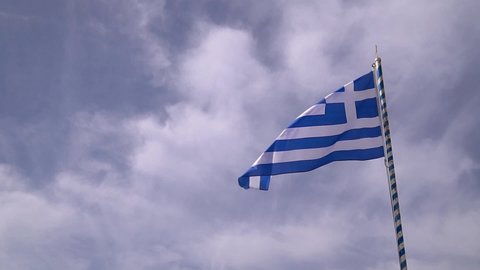 Greek national flag shot in slow motion flying in the breeze set against a blue sky background filled with white fluffy clouds