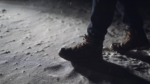 Close-up of male legs steps in winter snowy dark park. man walking in winter night. Black winter boots on legs. Snow falling in darkness. Guy In Snowy Weather At Cold Temperature Walking Alone night