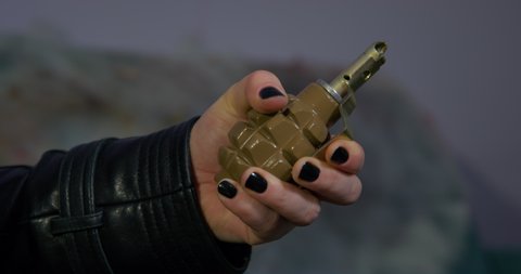 Criminal woman in leather jacket activates hand grenade on blurred background. Female hand with black nails holding dangerous projectile closeup
