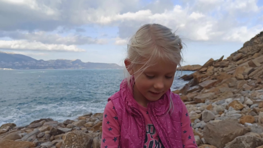 Portrait of little blonde girl singing a song enjoying nature sitting on the beach. Happy childhood.