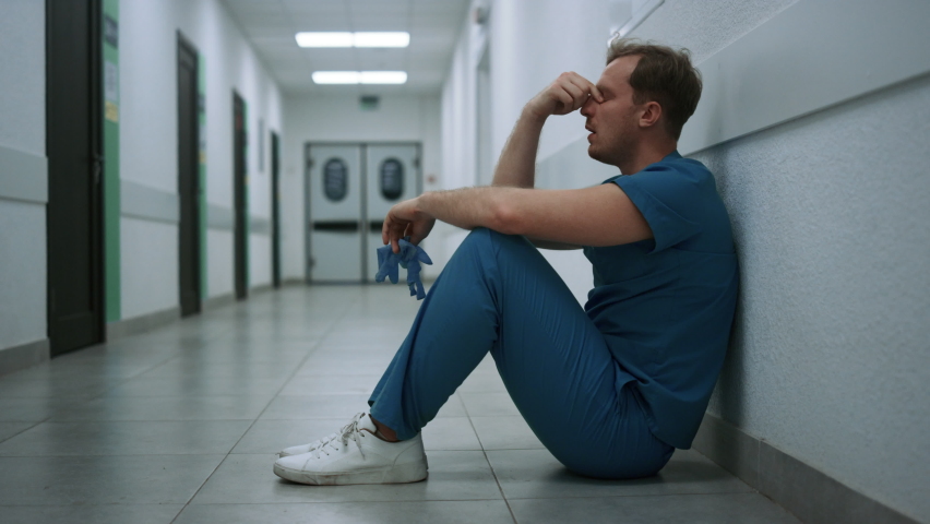 Stressed surgeon staying in empty clinic hallway after hard operation. Depressed doctor sitting alone on floor hospital corridor. Unhappy man physician throwing medical gloves holding hand on face. Royalty-Free Stock Footage #1090115751