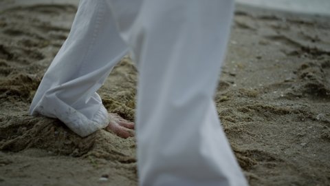 Closeup man feet taking steps on wet sand wearing white kimono. Unknown barefoot sportsman training on beach summer morning. View of dirty legs walking seacoast doing karate exercises. Sport concept.