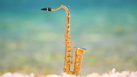Mini model of real alt tenor saxophone stands white small pebbles on seashore, against background of blue turquoise water. Music screensaver for romance. Copy space for your text. Beautiful picture.