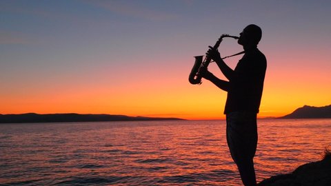Black Silhouette Young slender musician saxophonist with alt saxophone Stands on cliff, on seashore plays against backdrop of bright orange sunset, sky, sea waves, dawn, sea. Romantic music.