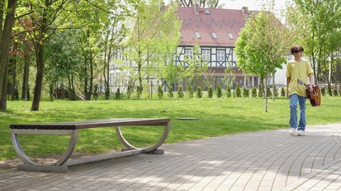 WROCLAW, POLAND - MAY 06, 2022: Teenager boy alone sit down on park bench play guitar boring and entertaining himself