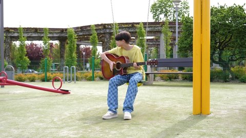 WROCLAW, POLAND - MAY 06, 2022: Teenager boy kid alone boring sit sway on swing play guitar on playground