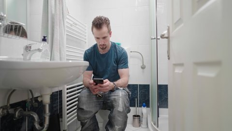 Opening door to bathroom with man on toilet using smartphone 4K