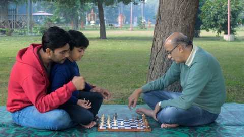 Grandfather, father, and a young grandson playing chess together - a board game, kids development, Indian family bonding . Family spending time together in a park - family bonding, friendly chess.