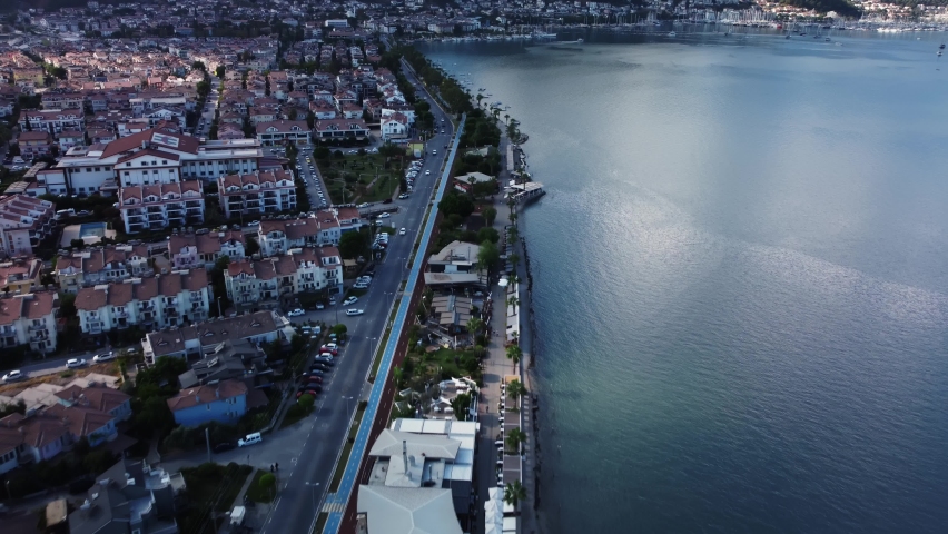 Fethiye city on the shore of Mediterranean sea with long and beautiful quay where tourists walk filmed by drone. Aerial view of the coastal city at warm summer day. Cars drive on the road along a pier