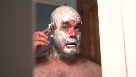 Rough, hairy man in front of the mirror with his face and head covered in shaving foam, shaving with a disposable blade.