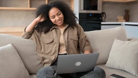 Smiling multiracial woman wearing domestic clothes sitting in the living room, relaxing and watching comedy show, looking at the laptop screen, staying connected during pandemic