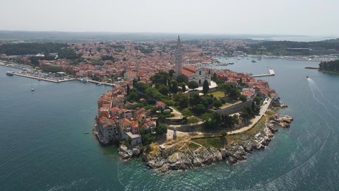 Aerial rotating backward shot of St. Euphemia church with the view of medieval old town in Rovinj in the background, Istria, Croatia surrounded by sea water at daytime.