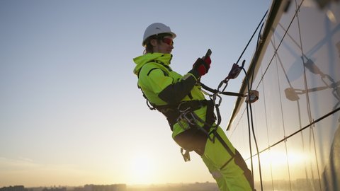Industrial climber reconciles received information via phone call. Brave climber hanging on safety ropes on skyscraper makes phone call at sunset