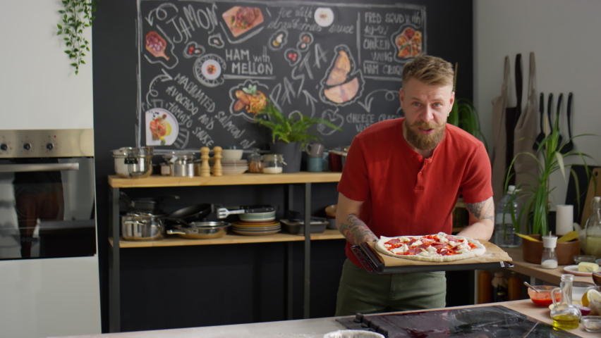 Bearded male food blogger looking at camera, speaking and then putting baking sheet with pizza into oven while giving online cooking class in kitchen