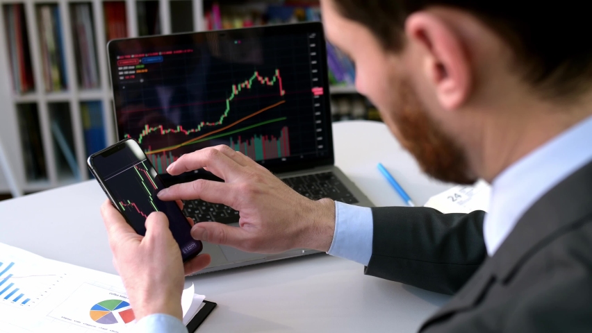 Exchange market. Smart man investor and crypto trader, using laptop and cellphone, analyzes charts of trading in stock market and digital cryptocurrency exchange,conducts analysis,trading crypto coins Royalty-Free Stock Footage #1090135815