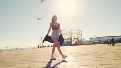 A bright stylish brunette in sunglasses in summer on the pier in denim shorts on the Santa Monica Pier at sunset. A positive sexy girl. The general plan is slow motion. A pretty woman enjoys life