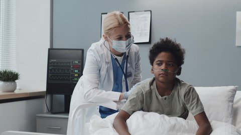 Medium long of mature Caucasian female doctor in medical mask examining Black boy with stethoscope, kid sitting on hospital bed at daytime