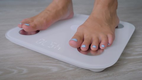 Female bare feet with pedicure stepping on white digital floor scales - woman weighing herself at home: close up, ground view. Measuring weight, control, wellness and diet concept