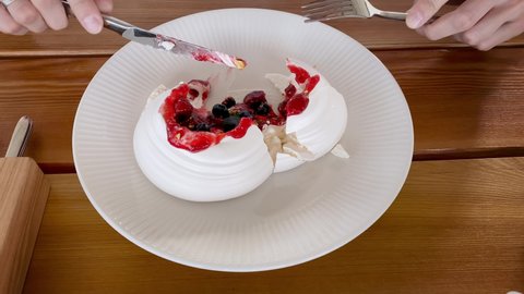 cake Anna Pavlova decorated with berries on white plate on wooden table. Confectioner cuts fresh baked pastry with knife and filling flows out closeup