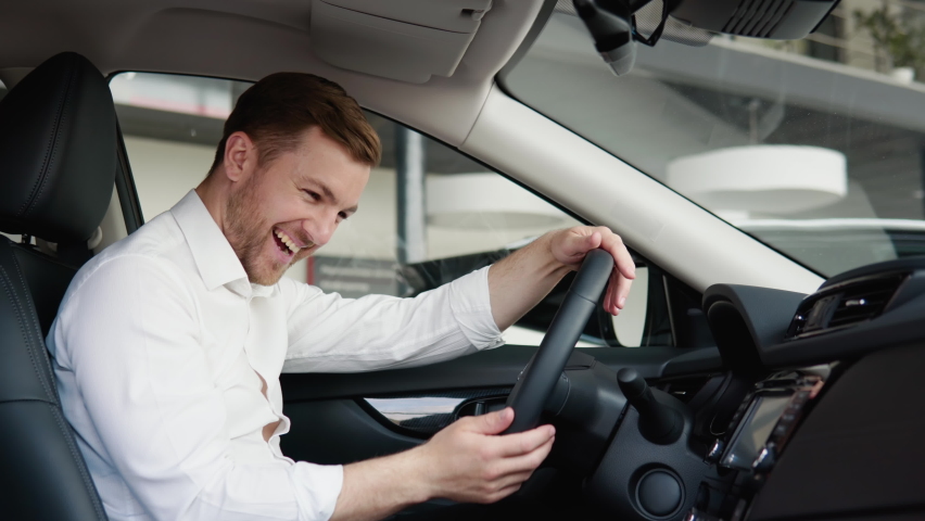 Portrait of a happy young man driving a new luxury electric car in a car dealership. The man shows emotions of happiness while driving in her new car. The buyer selects a car Royalty-Free Stock Footage #1090143377