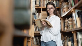 Young attractive student in glasses turns pages in the book, which she took from books on shelves in the library