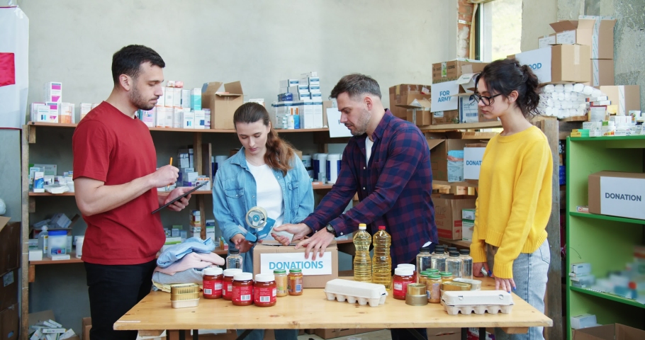 Group of hardworking volunteers in red cross charity center taping cardboard boxes with collected donations. Young people using digital tablet while preparing next batch of aid for sending.