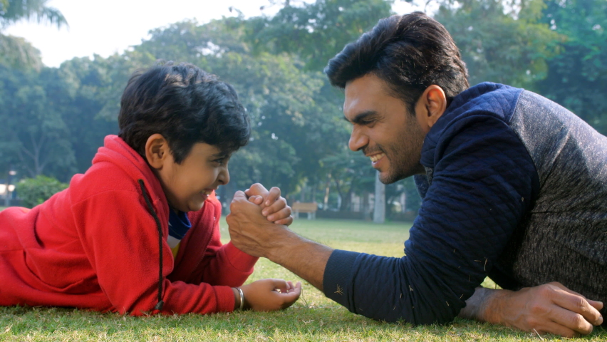 A young Indian boy doing arm-wrestling with his father - picnic, Indian family, physical strength, parenting. An excited little kid playing with his father in a park - father-son bonding, quality time