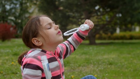 Cute little girl blows soap bubbles, plays in the park in summer. Child plays blowing beautiful bubbles, a happy child in the park. A lot of soap bubbles, the child experiences emotions of happiness.