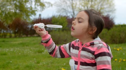 Cute preschooler girl blows soap bubbles, plays park in summer. Child plays blowing beautiful bubbles, a happy child in the park. Lot of soap bubbles, child experiences emotions of happiness.