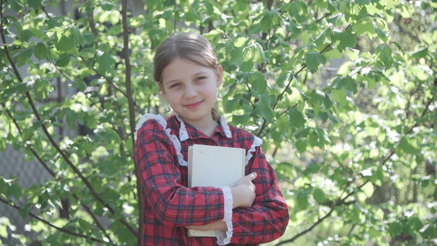 Portrait of a junior high school student. The girl is holding a textbook in her hands and smiling while looking at the camera. Sunny day, green background. Royalty-Free Stock Footage #1090150265