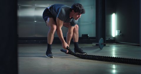 Cinematic shot of young fit virtual instructor or online personal trainer is showing web training workouts with battle ropes in sports studio gym.