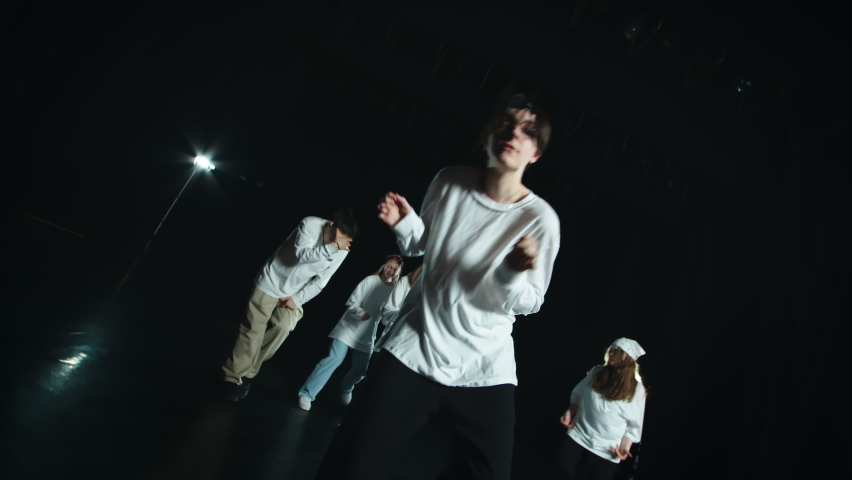 The dance team performs hip hop. All people are dressed in white. In the foreground is a guy with glasses and black hair. They move their bodies to the music, swing their arms and shake them