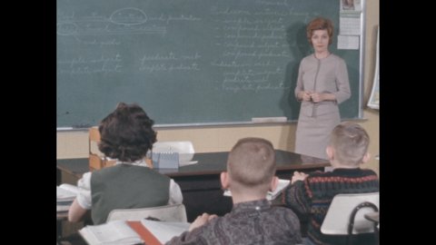 1960s: Woman holding clapperboard, clapping. Teacher lecturing to classroom. Students sitting at desks in classroom. Woman holding chalk, pointing to chalkboard.