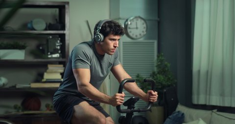 Cinematic shot of young fit man is putting on headphones with music for body workout and starts training at stationary bicycle at home.