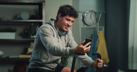 Cinematic shot of young fit smiling man is answering on video call from friend with smart phone while having training body workout at stationary bicycle at home.