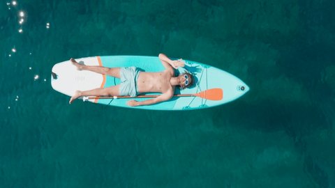 Directly above aerial shot: Man on vacation relaxes lying down on stand up paddle board in turquoise coloured tropical water. Man enjoying summer vacation 