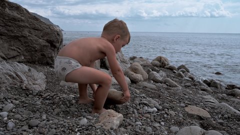 Cute baby boy playing on the seashore with rocks and pebbles. The kid is having a good time on a family vacation. Slow motion video