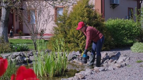 Adult Caucasian woman lets down a submersible water pump into an artificial garden fish pond to pump out water and clean the bottom from dirt and silt. Spring or Summer Pond Care