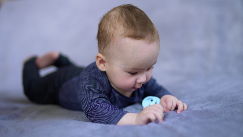 Cute little boy in blue shirt lies on belly and stares at his hands. Caucasian kid on the bed with pacifier near him.