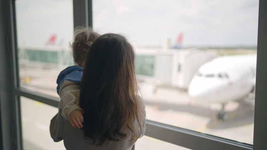 Close-up of attractive young mother and little daughter looking out window airport terminal, with airplane on background. Mother looks out an airplane window holding a baby. Toddler sleeping inside Royalty-Free Stock Footage #1090156477