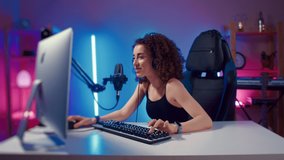 Young woman wearing headphones playing computer game neon fashion room, winner. Female gamer girl blogging, commenting hard match, looking at computer monitor, using computer mouse and keyboard