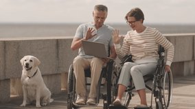 Modern wheelchair couple video chatting on laptop outdoors while their light yellow Labrador retriever resting nearby