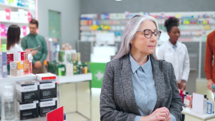 Portrait of mature caucasian woman standing near pharmacy showcase. Attractive senior woman looking and smiling at camera in modern drugstore with workers and clients on background.