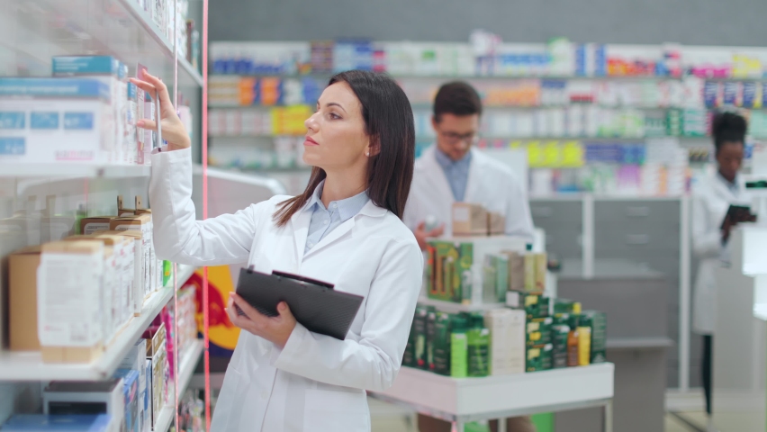Dark haired female apothecary in white lab coat writing on clipboard near drugstore showcase. Focused woman doing stocktake in modern pharmacy with working colleagues on background. Royalty-Free Stock Footage #1090170551