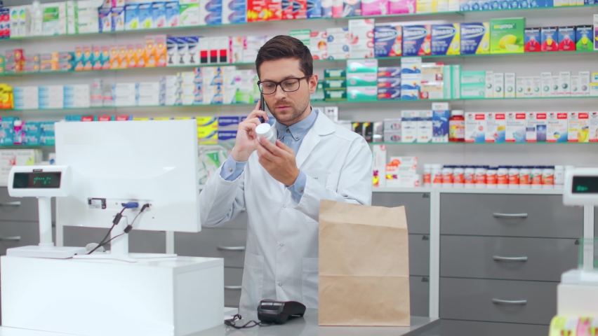 Male pharmacist in white lab coat and glasses having phone conversation with client while putting bottle of prescribed pills in bag. Caucasian man taking delivery order at modern drugstore.