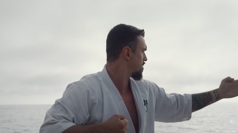 Serious athlete practicing karate in front gray cloudy sky close up. Tattooed bearded man workout fighting exercises wearing white kimono on beach. Focused sportsman making hands punches outdoors.