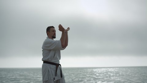 Taekwondo man training physical strength near calm sea. Bearded sportsman practicing martial arts wearing kimono overcast weather. Focused strong fighter making hands exercises workouting outdoors.