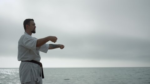 Bearded sportsman practicing martial arts standing beach gloomy morning. Caucasian man learning karate on cloudy nature outside. Serious athlete training stamina on seacoast. Sport activity concept.