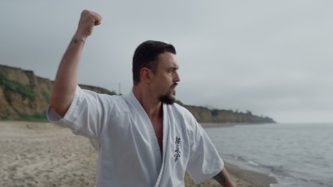 Athletic karate fighter exercising cloudy morning on sandy beach close up. Strong bearded man training martial exercises wearing kimono outdoors. Focused sportsman workout hands punches on nature.