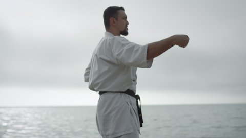 Confident man practicing hands position doing karate near calm sea. Bearded sportsman training fighting skills outdoors early morning. Strong focused athlete honing combat technique. Sport concept.